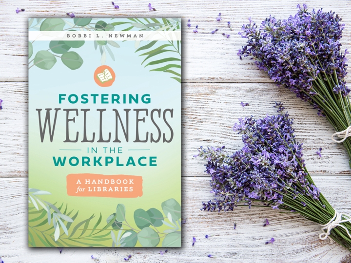 A copy of the book "Fostering Wellness in the Workplace" on distressed wood with some lavender next to it. 